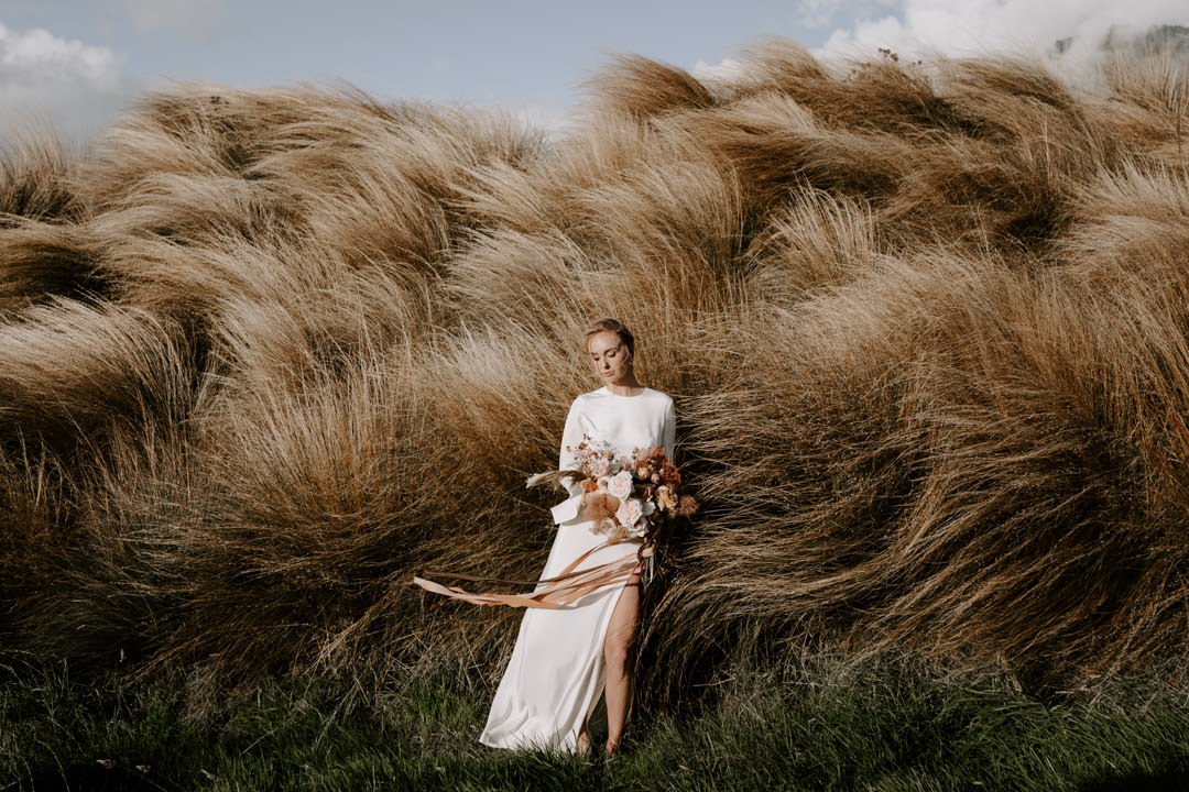 Bride wears Karen Walker Ritual Gown standing in tussocks at Jacks Retreat Elopement Wedding in Queenstown, New Zealand by Dawn Thomson Photography